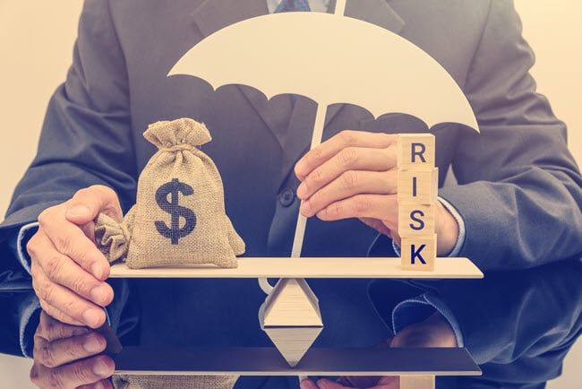 En person i kostym balanserar en pengasäck mot träklossar med ordet RISK under ett paraply.